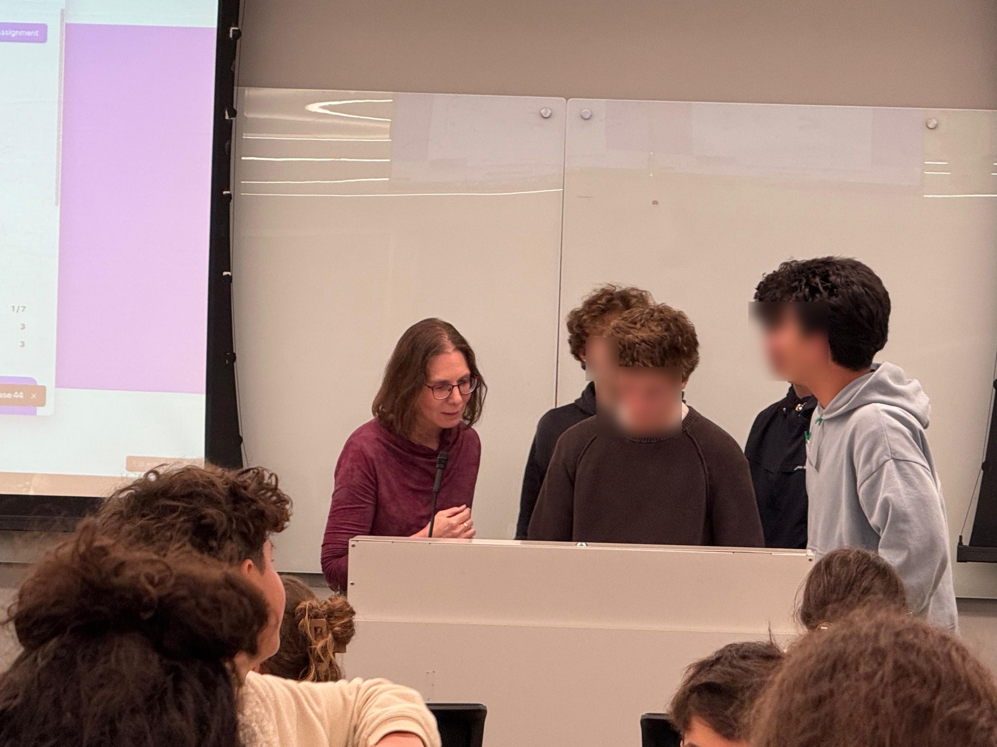 Adi speaking with teens at a podium during a hackathon session, presentation screen visible in the background